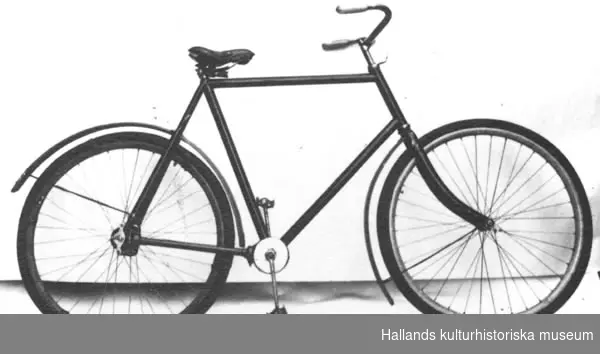 Cykel av stålrör. Diamond-ram. Spiralfjädrad, stoppad lädersadel. Styrhandtag av trä, målade gråröda. Träfälgar, oljestrukna, på suspensionsekrar. Bakdäcket provisoriskt säkrat till fälgen genom sex förankringar av isoleringsband. Sammansatt vev. Pedaler utan gummi. Kraftöverföring genom kardan, inkapslade vinkeldrev förmedlar kraften från vevaxeln till baknavet, vilken ej har frigång. Lika som föremålsnummer 029834. Ej frihjul. Svart. Styre, ring över gaffelkronan och vevparti förnicklade. Placeringen av skruvhål på styrrörets framsida antyder Crescent-märke.