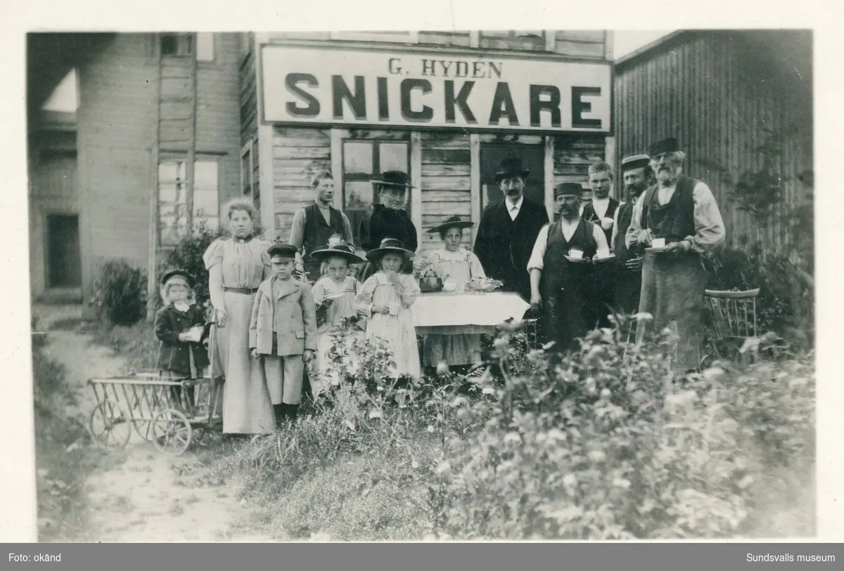 Gustaf Hydéns möbelsnickeri vid Bergsgatan i korsningen Thulegatan - Bergsgatan. Gustaf Hydén och hans fru (stadsbarnmorskan) Anna står i mitten bakom bordet. De fem barnen Rut, Gustaf, Anna, Mary och Elin står vid bordet. Den äldre flickan är troligen en barnflicka. Övriga personer är troligen anställda vid snickeriet.