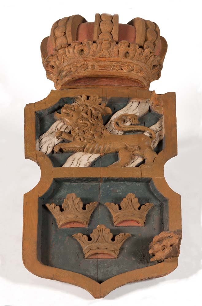 Akterspegelsornament, 2 delar. Dubbelkrönt stora riksvapnet i 2 längsgående hälfter med rester av lejontassar från sköldhållarna. Målad i gult, vitt, och rött på blå botten.