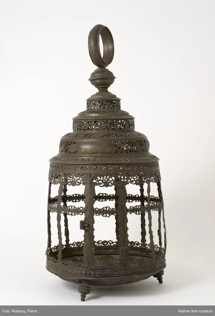 KLM 39725. Lykta. Av mässing. På tre fötter och med ring för upphängning. Med rikt genombrutet rankmönster. Driven mässingsplåt med bucklor på den trappstegsformade överdelen och på sidans nedre del. Tre knoppformiga fötter. I botten drivna bucklor samt en manlig och en kvinlig figur bland slingrande blad och blommor, en heltäckande dekor. Plats för 24 glasrutor, vilka saknas. Inuti tre ljushållare.