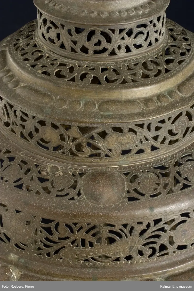 KLM 39725. Lykta. Av mässing. På tre fötter och med ring för upphängning. Med rikt genombrutet rankmönster. Driven mässingsplåt med bucklor på den trappstegsformade överdelen och på sidans nedre del. Tre knoppformiga fötter. I botten drivna bucklor samt en manlig och en kvinlig figur bland slingrande blad och blommor, en heltäckande dekor. Plats för 24 glasrutor, vilka saknas. Inuti tre ljushållare.