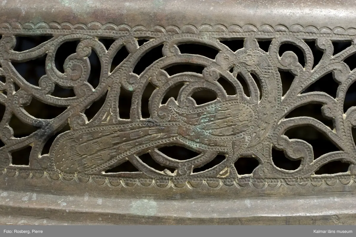 KLM 39725. Lykta. Av mässing. På tre fötter och med ring för upphängning. Med rikt genombrutet rankmönster. Driven mässingsplåt med bucklor på den trappstegsformade överdelen och på sidans nedre del. Tre knoppformiga fötter. I botten drivna bucklor samt en manlig och en kvinlig figur bland slingrande blad och blommor, en heltäckande dekor. Plats för 24 glasrutor, vilka saknas. Inuti tre ljushållare.