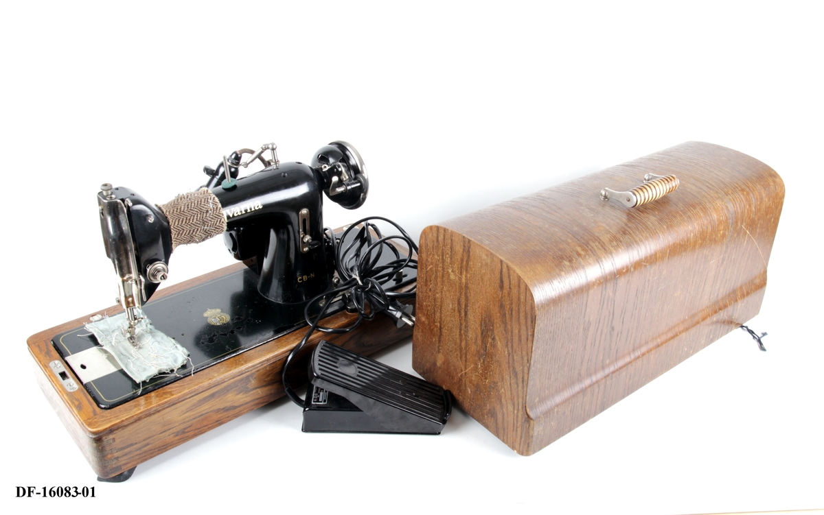 Symaskin på tresokkel, med tilhørende lokk med lås og håndtak. Symaskinen er utstyrt med lampe og  elektrisk motor, festet på baksiden med ledning og pedal.
