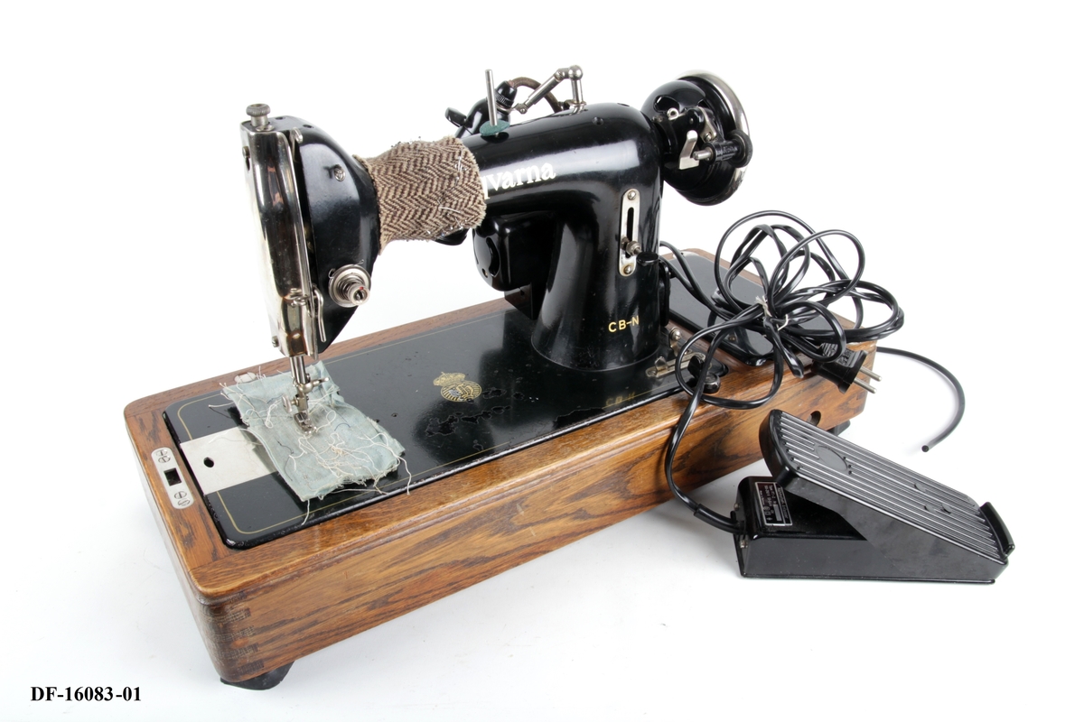 Symaskin på tresokkel, med tilhørende lokk med lås og håndtak. Symaskinen er utstyrt med lampe og  elektrisk motor, festet på baksiden med ledning og pedal.