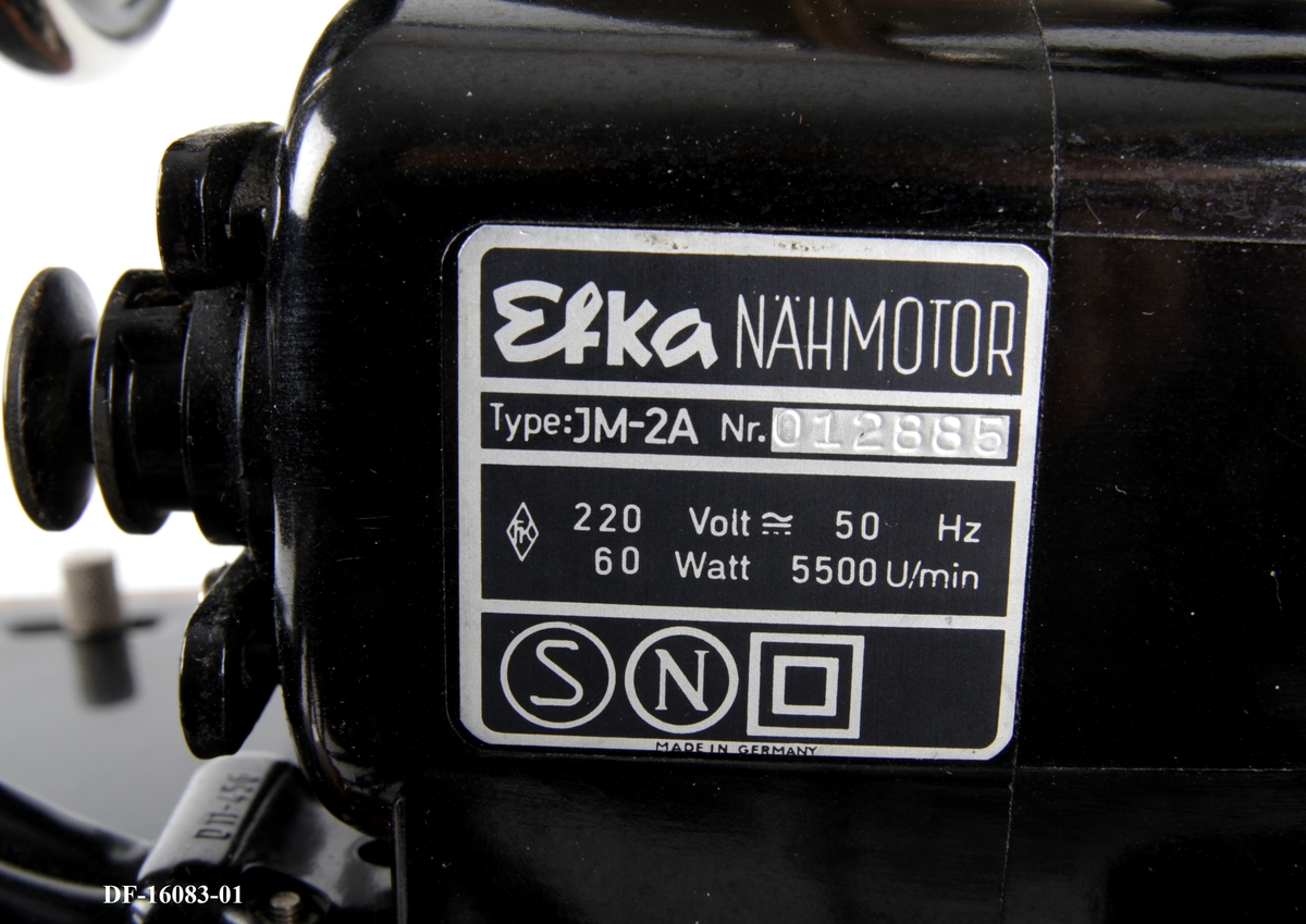 Symaskin på tresokkel, med tilhørende lokk med lås og håndtak. Symaskinen er utstyrt med lampe og  elektrisk motor, festet på baksiden med ledning og pedal.