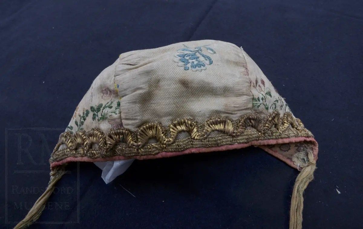 Dåpslue. Lys beige med broderier. Silke, diagonal kypert. Brodert med beige, blått rose og gulltråd.