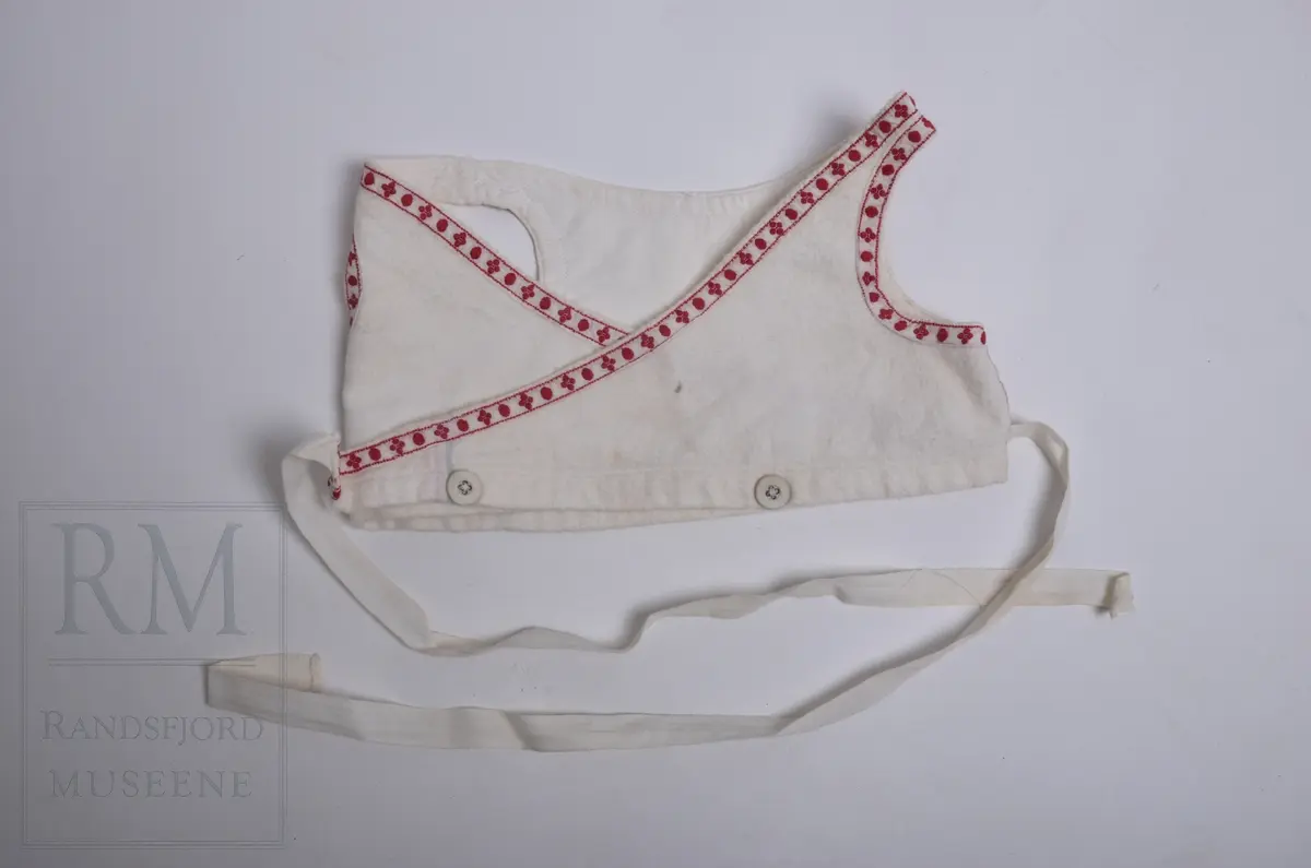 Liv til løyert e.l. til spebarn. Hvit flanell fóret med hvitt kypertvevet bomullstoff. Flanellen har lyse fargestriper langs kantene. Livet er kantet mot bomullsbpnd med rødt mønster. Omslagsliv med bendelbånd til å knytte med. 3 Lerretsknapper.