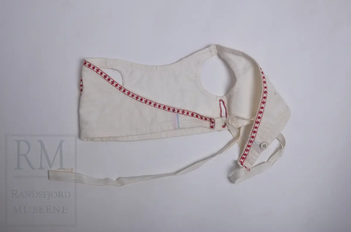 Liv til løyert e.l. til spebarn. Hvit flanell fóret med hvitt kypertvevet bomullstoff. Flanellen har lyse fargestriper langs kantene. Livet er kantet mot bomullsbpnd med rødt mønster. Omslagsliv med bendelbånd til å knytte med. 3 Lerretsknapper.