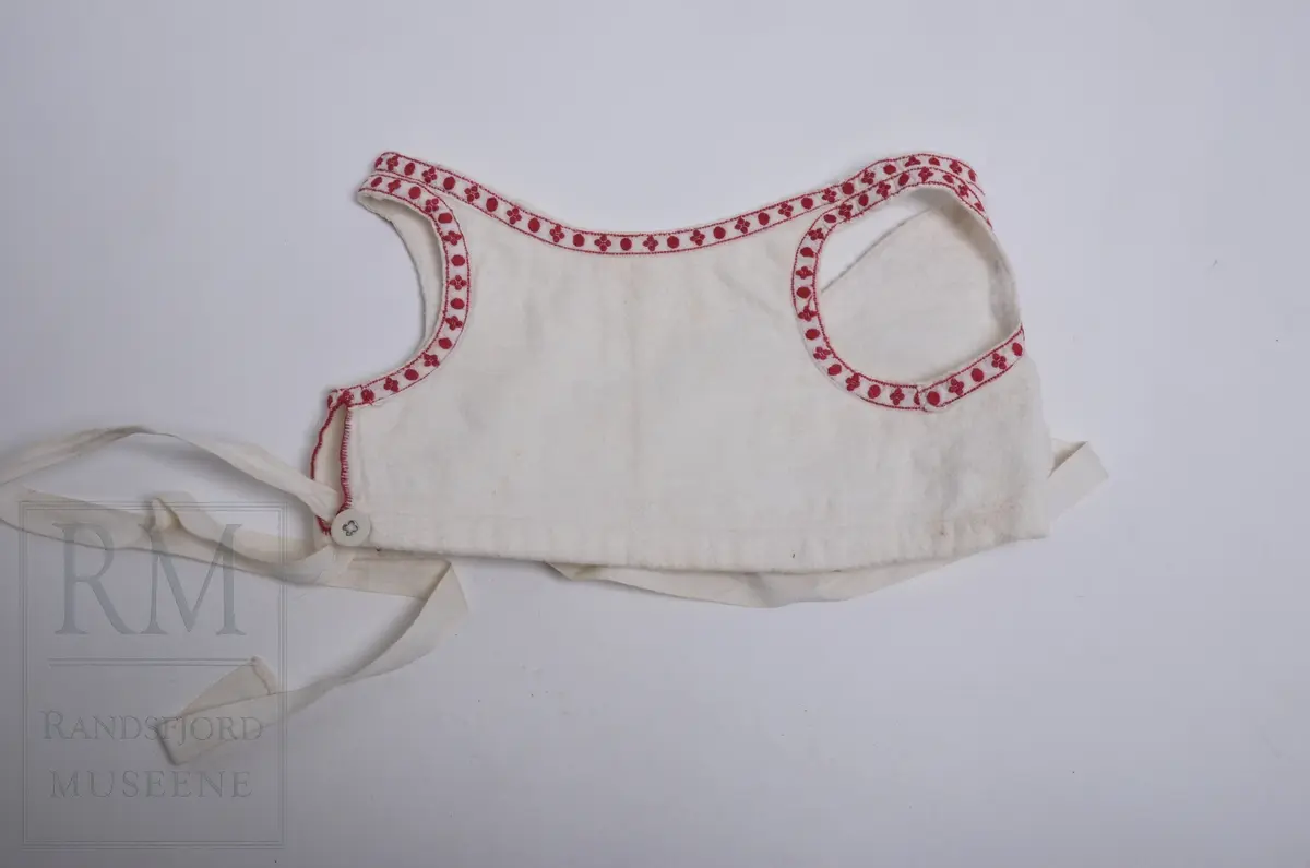 Liv til løyert e.l. til spebarn. Hvit flanell fóret med hvitt kypertvevet bomullstoff. Flanellen har lyse fargestriper langs kantene. Livet er kantet mot bomullsbpnd med rødt mønster. Omslagsliv med bendelbånd til å knytte med. 3 Lerretsknapper.