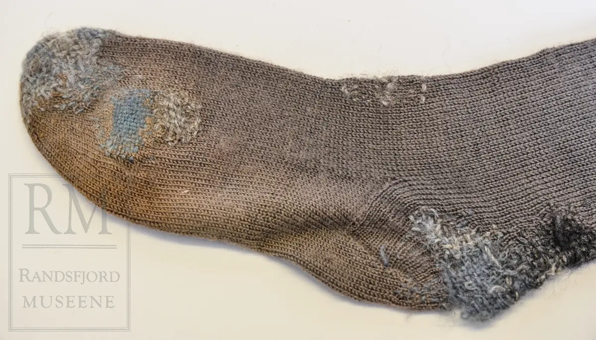 Strømper, 1 par, kvinne. Håndstrikket av ullgarn, grå 8,5cm vrangbord 2r2vr øverst. Kilehel og rund tåfelling. Felling bak på leggen. Innfelt strikket lapp på kneet på B. Hull og stopping. Misfarget føtter.