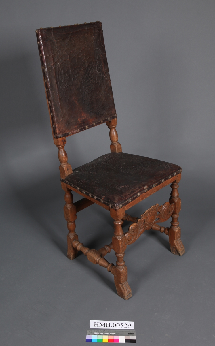 Stol med sete og rygg av lær og med dreiete buete bein. Mellom bein i front er det et utskåret stykke. Rosettformete nagler.