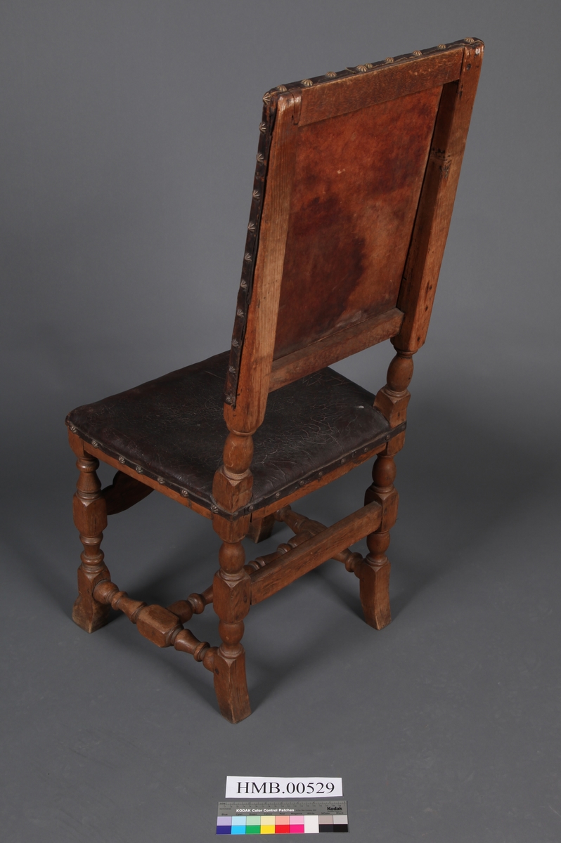 Stol med sete og rygg av lær og med dreiete buete bein. Mellom bein i front er det et utskåret stykke. Rosettformete nagler.