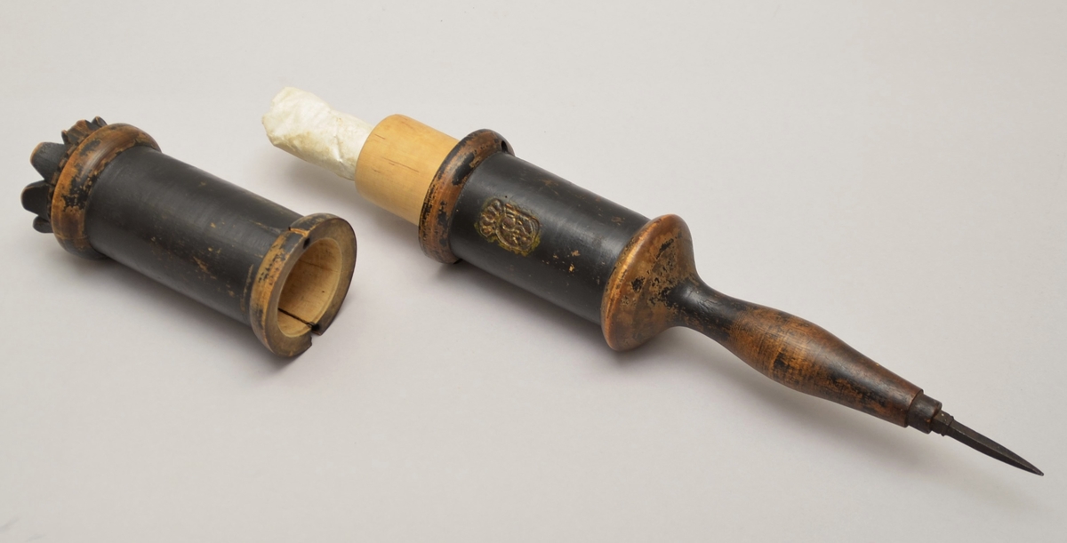 Bodstikke. Uthola sylinder i to delar.
Inni holrommet ligg eit samanrulla ark, papir er fest til eit tynt vove ark. Det er handskrive tekst på arket. Noko av byrjinga er borte, då noko av papiret er falt av der.
Sylindaren er i tre med ein jarnspiss i eine enda. Det er to små, gjennomgåand hol nedst på kvar del. Når dei to delane var sett saman, vart det sett inn ein pinne eller liknande gjennom hola på kvar side, slik at delane vart låste saman.
På eine sida og på enda er det eit symbol stansa inn i treet. Symbolet ser ut som ein stor C med noko inni. Over C-forma er ei krune.