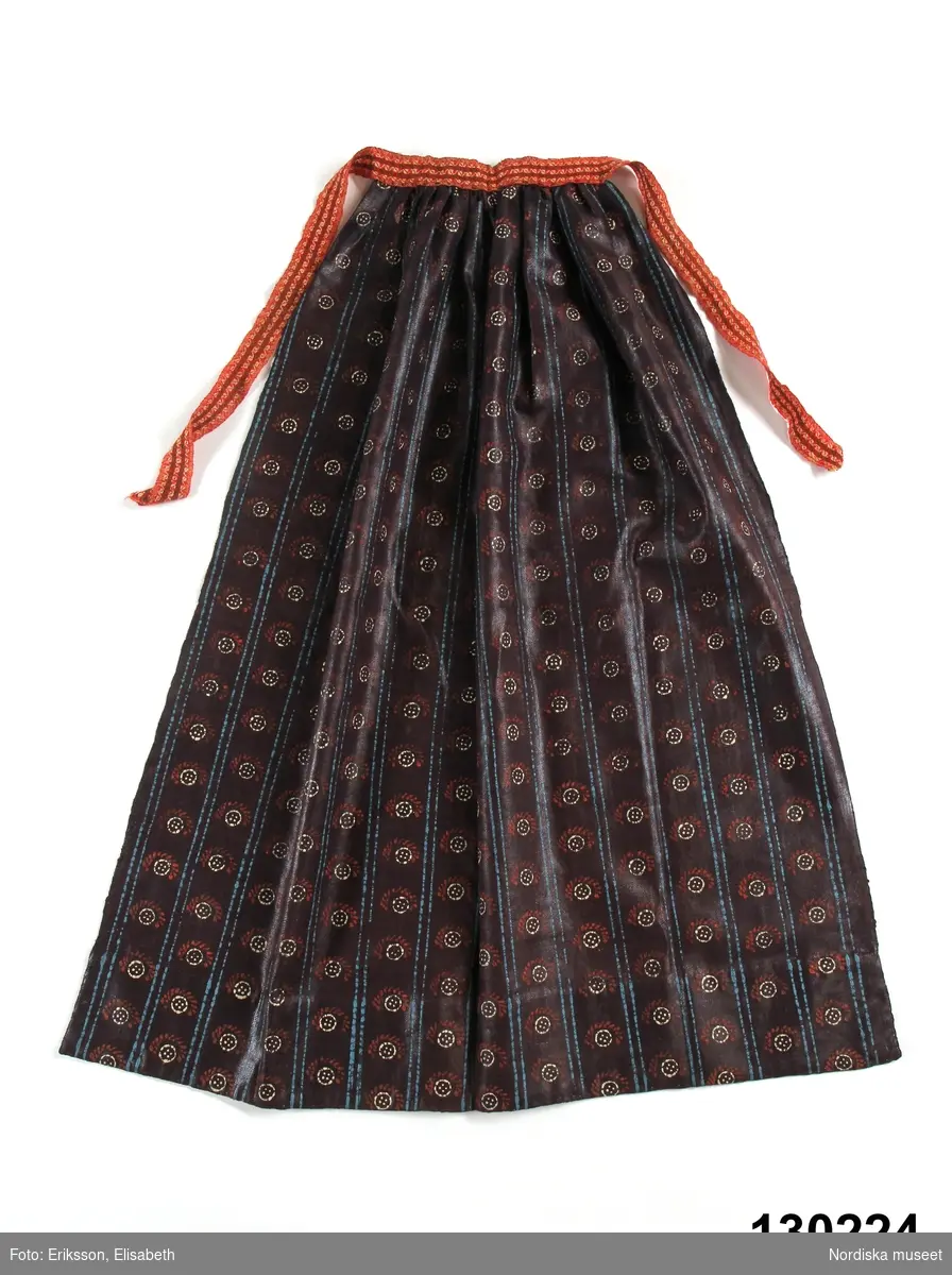 Förkläde av mörklila tunn yllekypert av kamgarn med starkt glättad yta, s.k. rask eller chalon. har fått ett sekundärt stämpeltryckt mönster med oljefärg, smala dubbla  ränder  i klarblått och mellan ränderna rader av  "rosor" dvs. mönsterfigurer i vitt och rött. Rynkat i midjan med ett påsytt  3 cm brett midjeband i ylle smårutigt  i rött, grönt och vitt av den sort som kallas Malsband dvs. Malungsband. Nederkanten inskodd med  smalrandigt halvylletyg i grått och grönt/ljusgult. Handsömnad.
Rask eller chalon var ett köpetyg som användes till högtidsförkläden. Här har man låtit en lokal tryckare mönstra tyget, vilket är ovanligt på denna typ av förkläde. 
erit Eldvik 2012-03-29