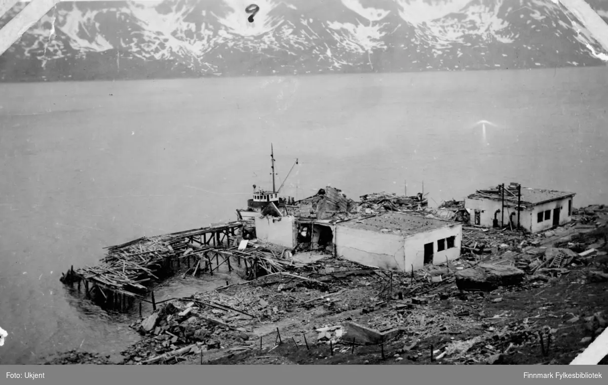 Ruiner av et fryseri i Øksfjord etter ødeleggelsene 1944. Man kan se deler av en gammel kai, og rester etter bygninger og andre konstruksjoner på bakken. Ved siden av kaia ligger det en liten båt. På fiskeriannlegget kan man se dør- og vindusåpningene der vinduene og dørene en gang var.