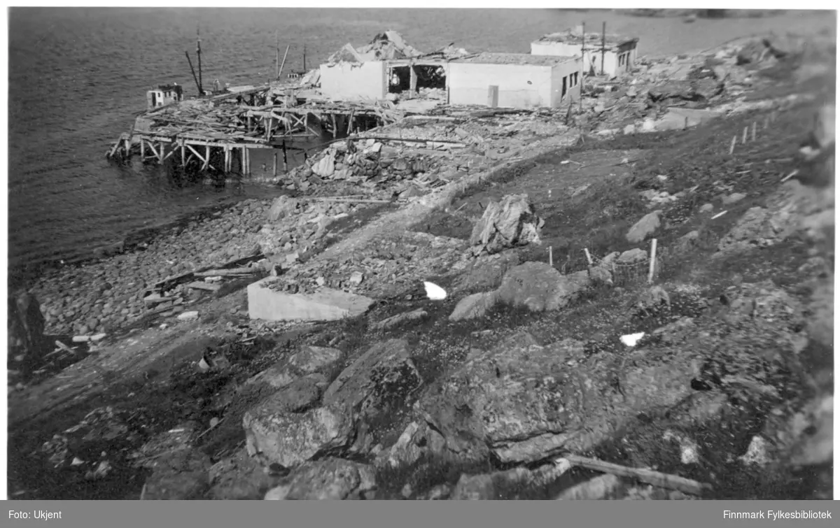 Ruiner av et fryseri i Øksfjord etter ødeleggelsene 1944. Man kan se deler av en gammel kai, og rester etter bygninger og andre konstruksjoner på bakken. Ved siden av kaia ligger det en liten båt. På fiskeriannlegget kan man se dør- og vindusåpningene der vinduene og dørene en gang var.