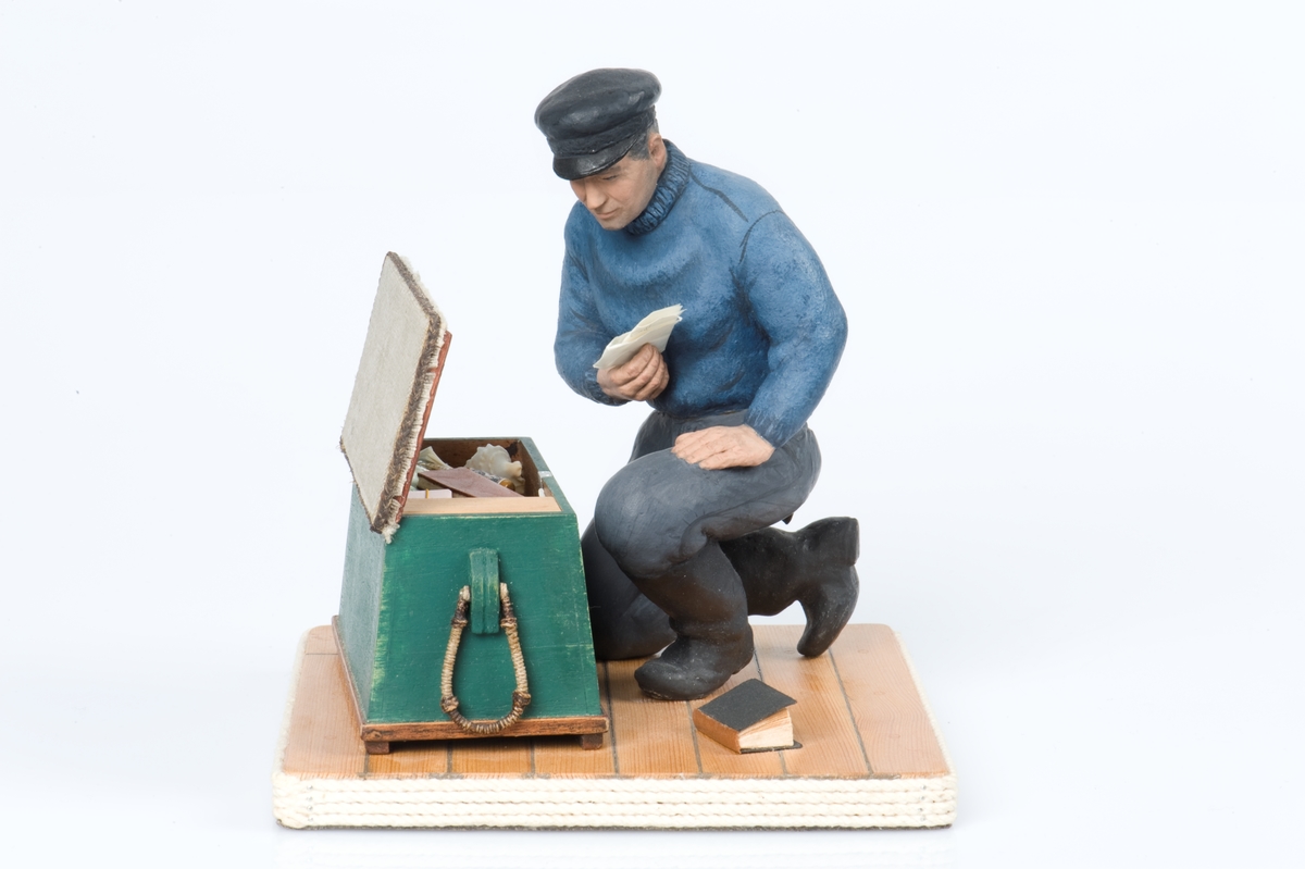 keramikfigur av en sjöman vid sin sjömanskista. Flera små detaljer i kistan. Bl a fartygsporträtt av barkskepp, målat av Hendel Friberg.