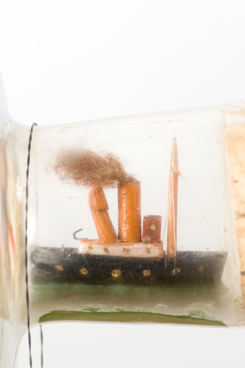 Flaskskepp med femmastad fullriggare. Seglen är beslagna och i flaskans hals är en liten bogserbåt behjälplig. I bakgrunden är en bebyggd strand. Tillverkaren är okänd.