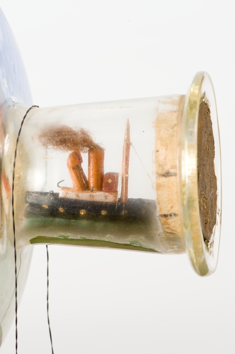 Flaskskepp med femmastad fullriggare. Seglen är beslagna och i flaskans hals är en liten bogserbåt behjälplig. I bakgrunden är en bebyggd strand. Tillverkaren är okänd.