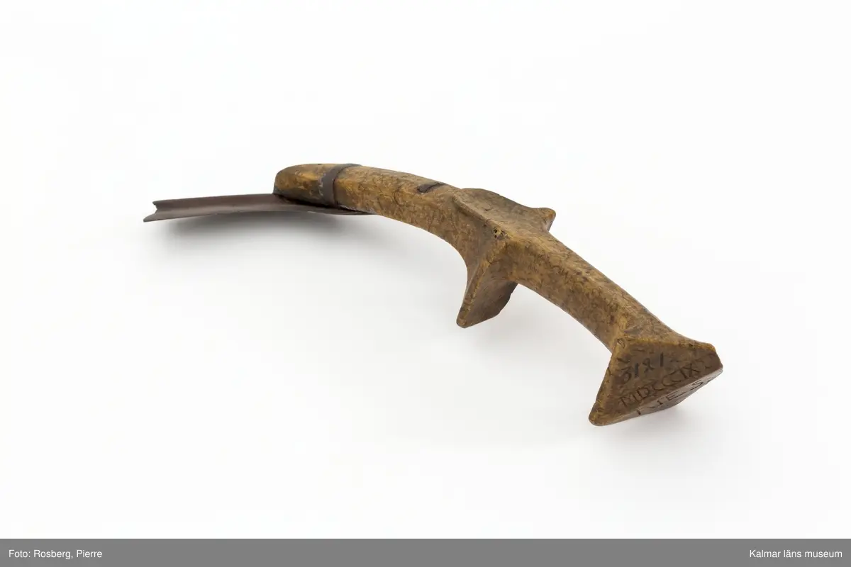 KLM 3121. Skära, vinkelskära, trä, metall. Vinkelskära med bågböjt blad av järn, avbrutet, fastnitat på skaftets ena sida. Skaft av masurbjörk med utskuret handtag och fyrkantig knopp, upphöjd på mitten. Bladet är målat i rödbrun färg under senare tid. På knoppens undersida inskuret monogramet N.E.S. och årtalet MDCCCIX (1809). Nära bladet brännstämplat PNS. Bladet märkt A. Användes till att skära av säden.