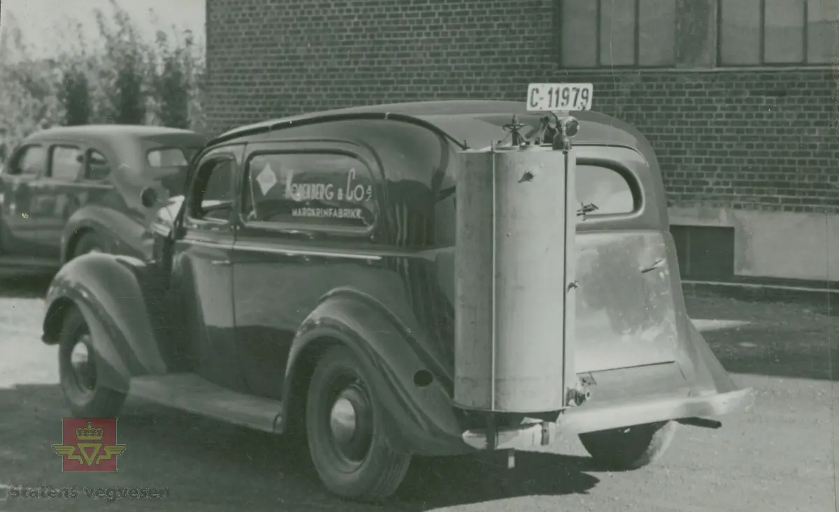 "Karbidgenerator produsert av Øveråsens Motorfabrikk, Gjøvik. De ble montert av Colbjørnsens & Co. A/S under hele krigstiden." I følge merking i album. På bilen med kjennetegn C-12178 står det skrevet "A/S Norske Konfektionsfabriker." På skiltet øverst på murbygningen står sannsynligvis "A/S Sveiseindustri." 

Bilde 1/4 er en Chevrolet med en eldre Chev. baklykt  på taket. 20.08.2016: "Bilde 1 er en Chevrolet 1939-40 varebil med norskbygd karosseri. "25.06.2018: Til venstre snuten av en Packard fra sent 1930-tall. Opplysninger fra Ivar Erlend Stav.
Klikk på pilen til høyre:
Bilde 2/4 er en Dodge 1935-1936 modell. (Strømmenbygd?)
Bilde 3/4  er en 1930 modell Brockway. 25.06.2018: Bildet 3 viser snuten av en Plymouth1937. Opplysninger fra Ivar Erlend Stav.
Bilde 4/4 er en Ford fra 1937-1938 modell." 25.06.2018:  "Ford 1938, siden det var første år med langsgående blanklist hos Ford. Karosseriet er originalt amerikansk varebilkarosseri på personbilchassis ("sedan delivery" - i motsetning til et mer grovbygd varekarosseri på lett lastebilchassis og lastebilfront, kalt "panel delivery". Opplysninger fra Ivar Erlend Stav. 

17.08.2016: Opplysninger til kjøretøyene på bildet v/tidligere daglig leder John Hindklev ved Norsk Kjøretøyhistorisk Museum. 

"Under andre verdenskrig besto virksomheten til firmaet Colbjørnsen stort sett av å montere og vedlikeholde karbidgeneratorer fra Øveråsen. Legg merke til hvordan man felte generatorene inn i varebilkarosseriene. Reklameteksten  ble delvis borte." Sitat fra boka: "Familiefirmaet-utenom allfarvei/2000 årsboken." Colbjørnsen & Co A/S, av Bjørn Ausjen Johannessen.