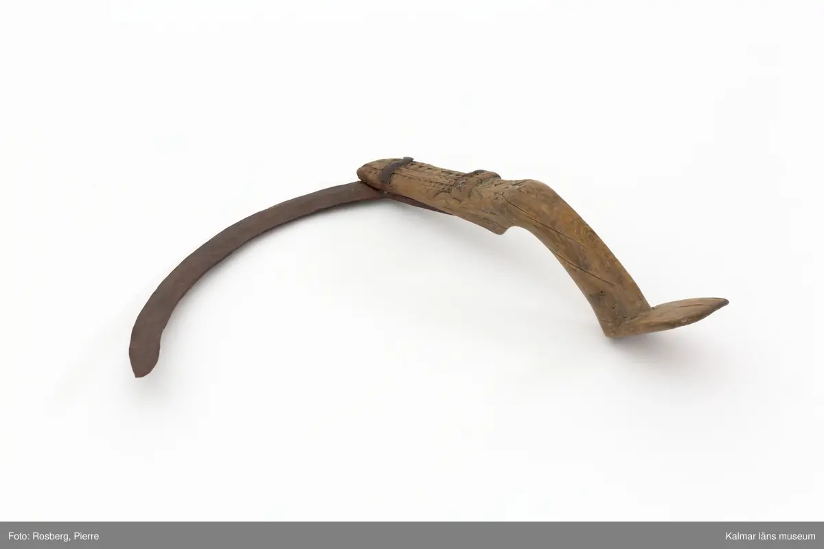 KLM 22639. Skära, vinkelskära. Av trä och metall. Vinkelskära med bågböjt blad av järn, fastnitat på skaftets ena sida. Skaft av trä med utskuret handtag och ovalformad platta i änden. Handtaget har skuren dekor i form av sicksack-mönster. Under skaftet en blomma med ring runt samt "LS" (= Lisa Svensson) ANO 1751. Bladet är målat i rödbrun färg, för rostskydd, under senare tid.