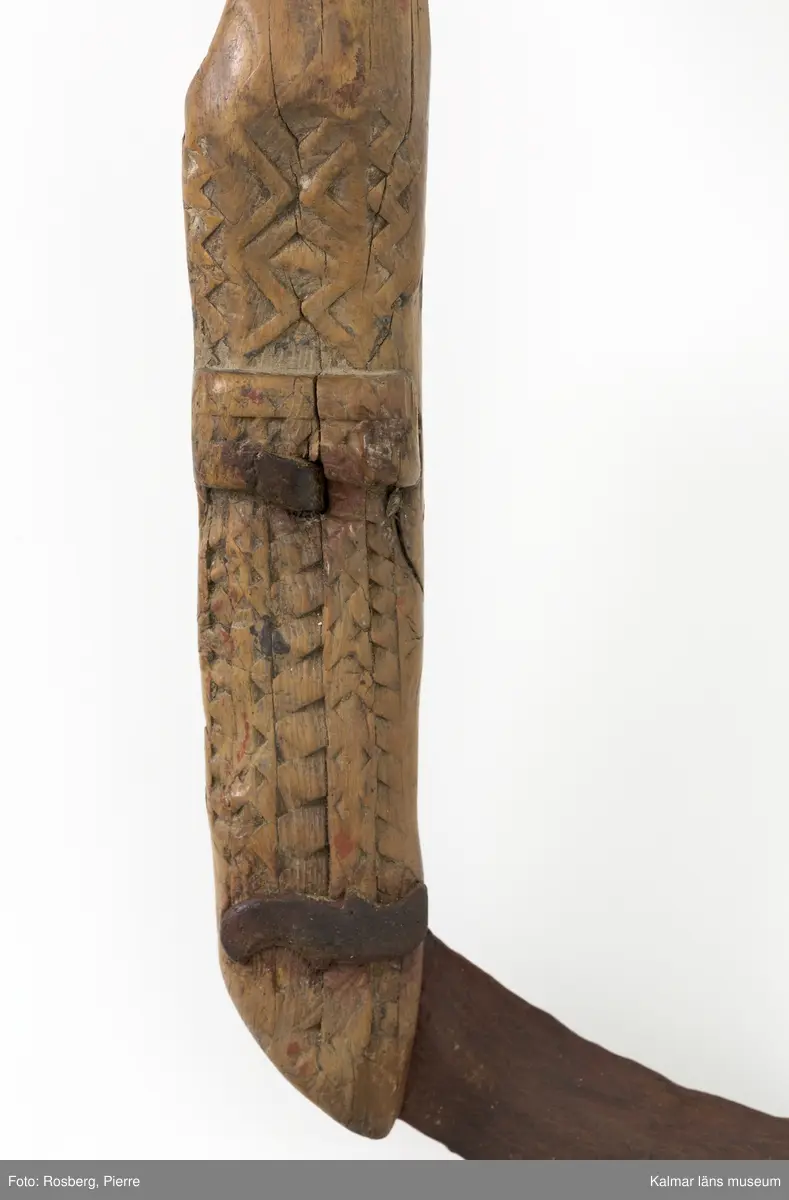 KLM 22639. Skära, vinkelskära. Av trä och metall. Vinkelskära med bågböjt blad av järn, fastnitat på skaftets ena sida. Skaft av trä med utskuret handtag och ovalformad platta i änden. Handtaget har skuren dekor i form av sicksack-mönster. Under skaftet en blomma med ring runt samt "LS" (= Lisa Svensson) ANO 1751. Bladet är målat i rödbrun färg, för rostskydd, under senare tid.