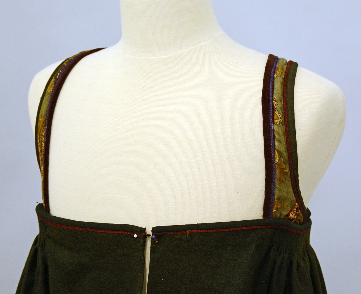 Stakk med påsydd liv. Skjørtet er rynket oppe, metallhekter på hver side av splitten/åpningen foran. Stoffet er av svart vadmel i lerretsbinding. Brodert mørkerød kant øverst på stakken. Skoningen er påsydd med et annet tøy enn det originale, den er 10 cm og har striper av mørkegrønn og sort fløyel.Skoningen er 10 cm. Nest nederste kant er mørkerød.Livet har kjøpetøy i trykteblomster med påsydde bånd. Halsutringningen er av mørkerødt fløyel med broderibord i lilla. Ermutringningene er av sort bomullstøy med mørkerød broderibord. Fòret er av rutet bomullstøy. Stakketøyet er falmet, har noen flekker,og små hull etter møll.