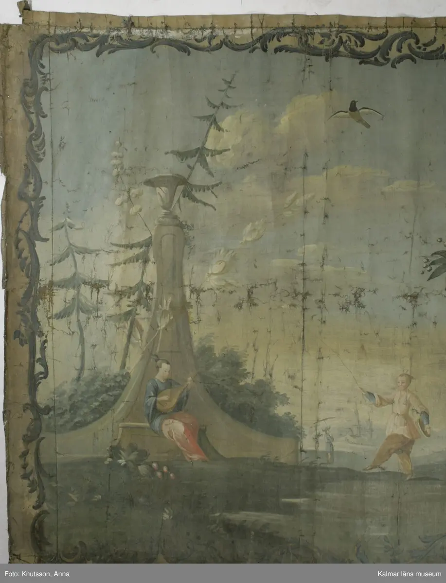 KLM 43973. Tapet, väggfält. Oljemålning på väv. Blå färg utanför ramen. Ramen är snirkliga rankor i mörkblått. Orientaliskt motiv med fem personer i orientaliska kläder. Första personen är en kvinna som spelar ett lutaliknande instrument. Kvinnan har blå överdel och byxor i en lysande röd färg. Nästa person har en ljusgul överdel och beige byxor. Det ser ut som om personen har en flygande fågel fastknuten i ett snöre. I den andra handen håller personen en slags platta. Nästa person sitter ner och håller en likadan platta. Längst till höger i bild står en kvinna böjd över en urna. Kvinnan har rosa överdel, blå byxor och lysande röda skor. I omgivningarna finns flera byggnader, tunna träd, växter och även blommor.