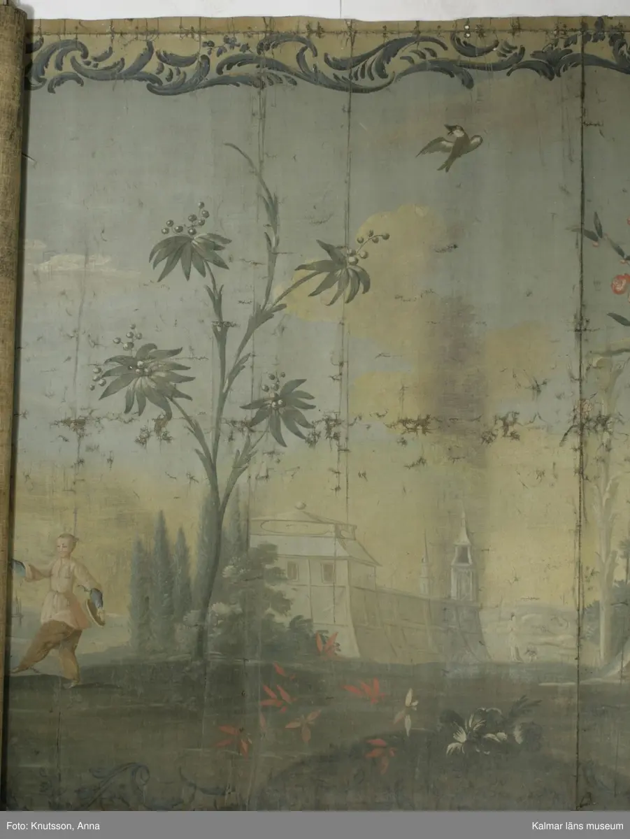 KLM 43973. Tapet, väggfält. Oljemålning på väv. Blå färg utanför ramen. Ramen är snirkliga rankor i mörkblått. Orientaliskt motiv med fem personer i orientaliska kläder. Första personen är en kvinna som spelar ett lutaliknande instrument. Kvinnan har blå överdel och byxor i en lysande röd färg. Nästa person har en ljusgul överdel och beige byxor. Det ser ut som om personen har en flygande fågel fastknuten i ett snöre. I den andra handen håller personen en slags platta. Nästa person sitter ner och håller en likadan platta. Längst till höger i bild står en kvinna böjd över en urna. Kvinnan har rosa överdel, blå byxor och lysande röda skor. I omgivningarna finns flera byggnader, tunna träd, växter och även blommor.
