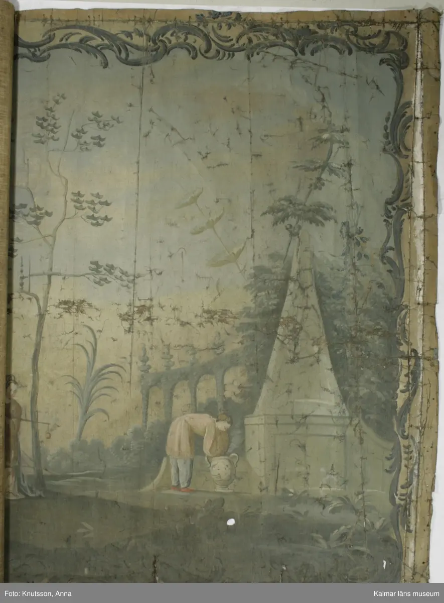 KLM 43973. Tapet, väggfält. Oljemålning på väv. Blå färg utanför ramen. Ramen är snirkliga rankor i mörkblått. Orientaliskt motiv med fem personer i orientaliska kläder. Första personen är en kvinna som spelar ett lutaliknande instrument. Kvinnan har blå överdel och byxor i en lysande röd färg. Nästa person har en ljusgul överdel och beige byxor. Det ser ut som om personen har en flygande fågel fastknuten i ett snöre. I den andra handen håller personen en slags platta. Nästa person sitter ner och håller en likadan platta. Längst till höger i bild står en kvinna böjd över en urna. Kvinnan har rosa överdel, blå byxor och lysande röda skor. I omgivningarna finns flera byggnader, tunna träd, växter och även blommor.