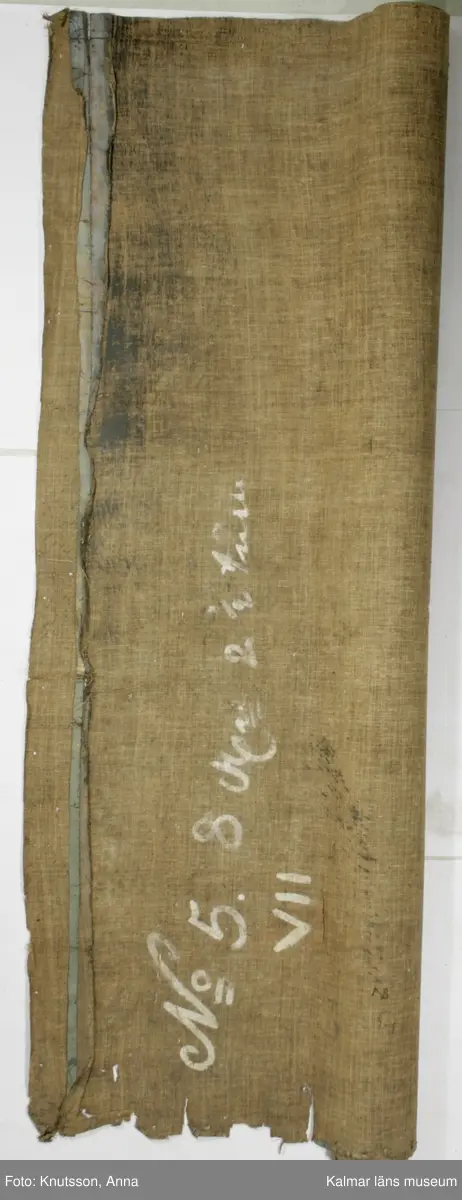 KLM 43973. Tapet, väggfält. Oljemålning på väv. Blå färg utanför ramen. Ramen är snirkliga rankor i mörkblått. Orientaliskt motiv med fem personer i orientaliska kläder. Första personen är en kvinna som spelar ett lutaliknande instrument. Kvinnan har blå överdel och byxor i en lysande röd färg. Nästa person har en ljusgul överdel och beige byxor. Det ser ut som om personen har en flygande fågel fastknuten i ett snöre. I den andra handen håller personen en slags platta. Nästa person sitter ner och håller en likadan platta. Längst till höger i bild står en kvinna böjd över en urna. Kvinnan har rosa överdel, blå byxor och lysande röda skor. I omgivningarna finns flera byggnader, tunna träd, växter och även blommor.