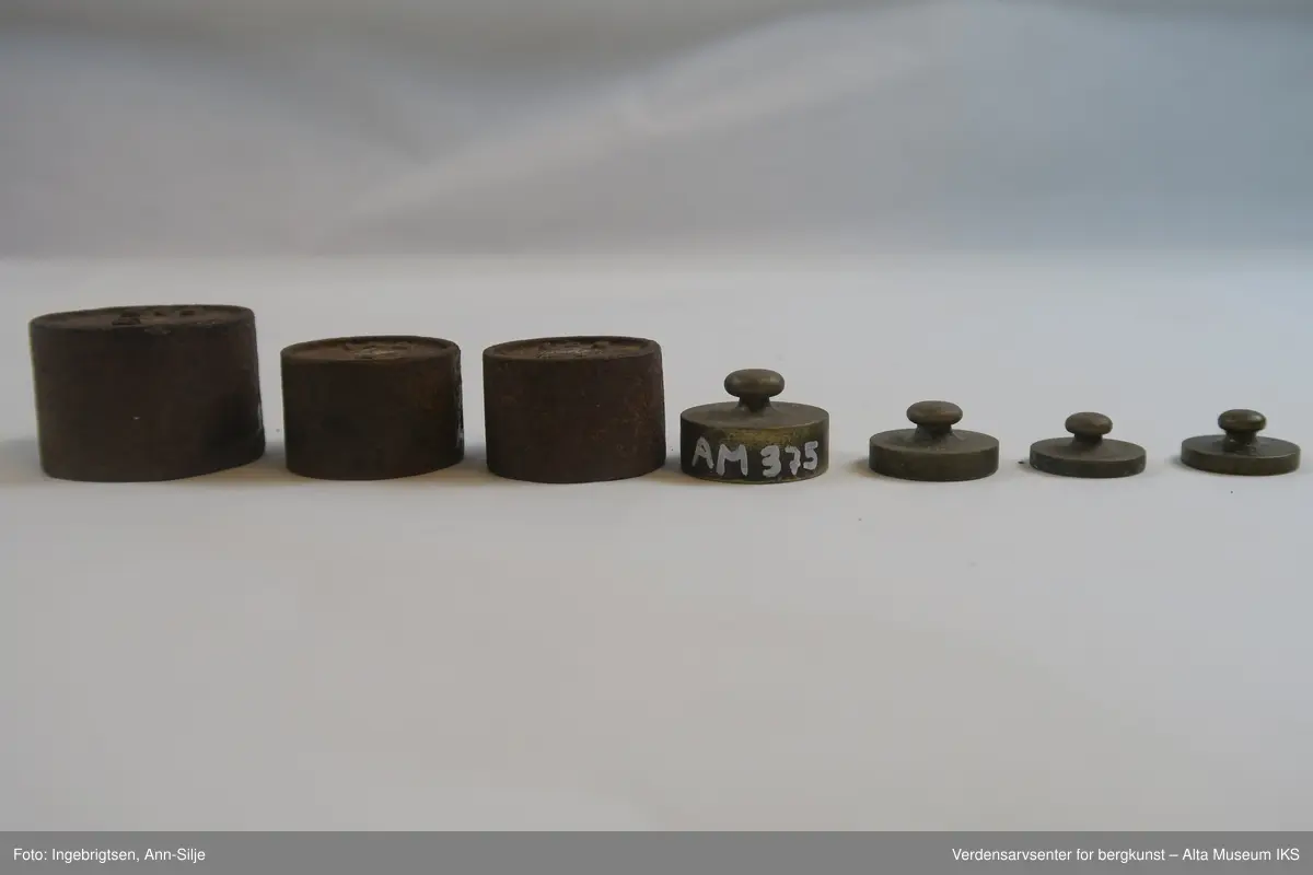 Rektangulær trebit, med hull tilpasset ulike loddstørrelser. Det er 7 lodd: 2 stk. 10 gram, 1 stk. 20 gram, 1 st. 50 gram, 2 stk. 100 gram og 1 stk. 200 gram. Alle loddene er sylinderformet med knott på toppen. Loddene varierer i størrelse: de minste er 2 cm i diameter og 1 cm i høyde, mens den største er 4 cm i diameter og 2,8 cm i høyde.
