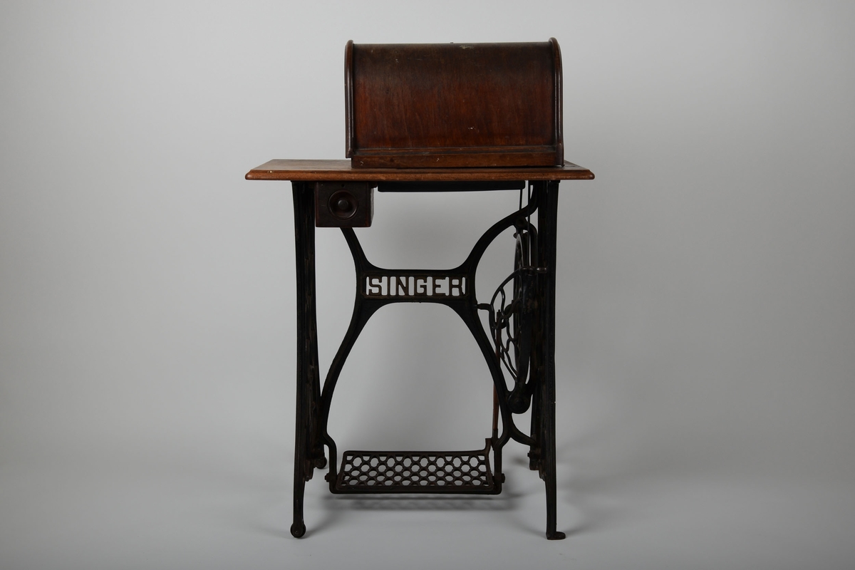 Symaskin med bord og lokk. Understell av støpejern, med pedal.

Treflaten som symaskinen står på har innfelte hull til å sette lokket i. En liten trebit står i et hull i bordet.