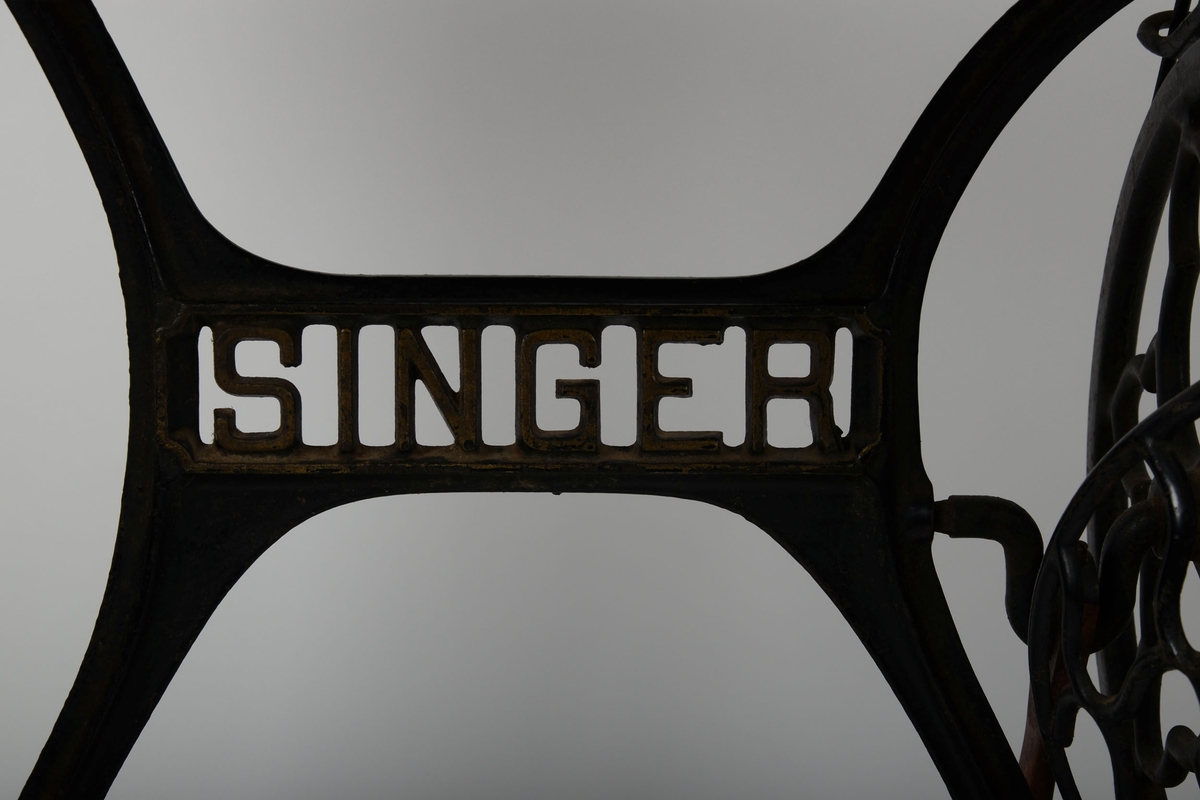 Symaskin med bord og lokk. Understell av støpejern, med pedal.

Treflaten som symaskinen står på har innfelte hull til å sette lokket i. En liten trebit står i et hull i bordet.