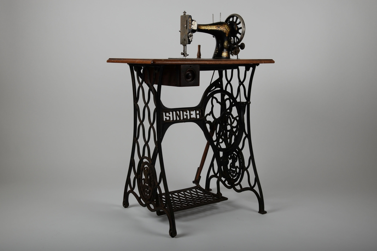 Symaskin med bord og lokk. Understell av støpejern, med pedal.

Treflaten som symaskinen står på har innfelte hull til å sette lokket i. En liten trebit står i et hull i bordet.