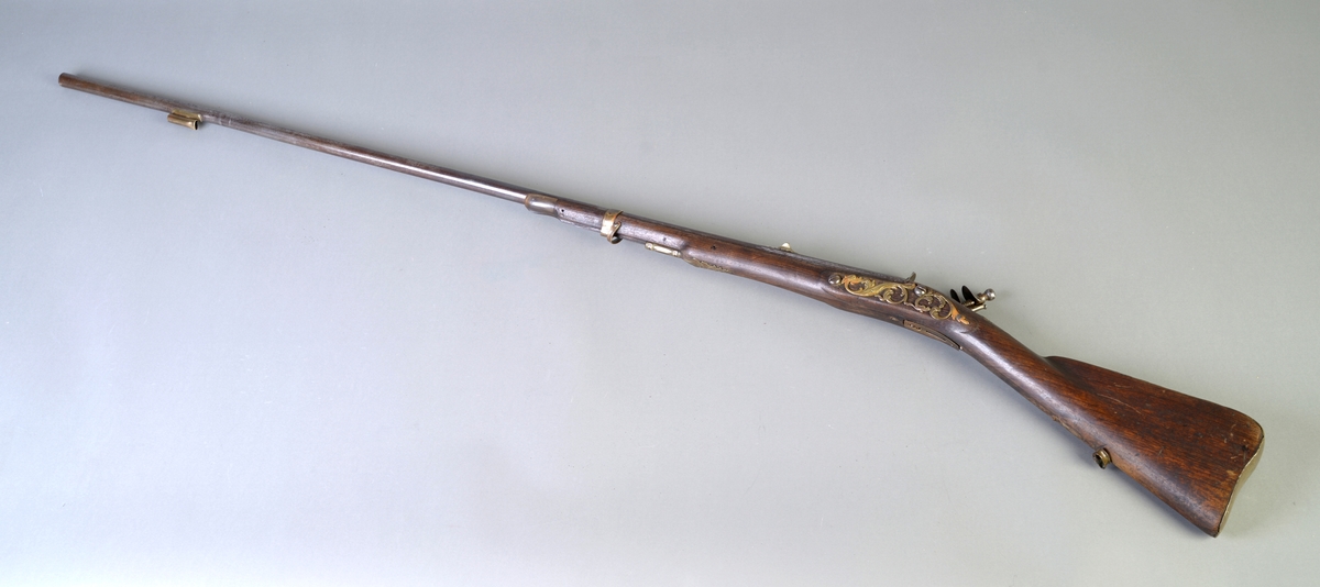 Flintlås og langt løp. På venstre side av låsen dekorativt messingbeslag, formet som en ranke. Hanen og beslaget ved denne av jern og prydet med rokokko ornamentikk. Messingbeslag ved avtrekkeren og på kolben. Ladestokken mangler.