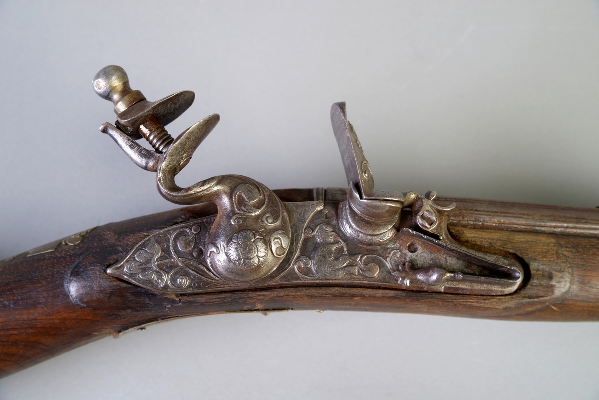 Flintlås og langt løp. På venstre side av låsen dekorativt messingbeslag, formet som en ranke. Hanen og beslaget ved denne av jern og prydet med rokokko ornamentikk. Messingbeslag ved avtrekkeren og på kolben. Ladestokken mangler.