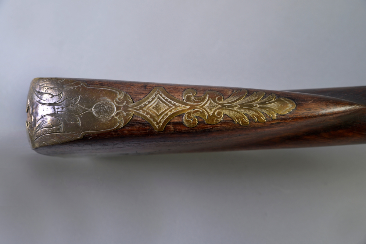Flintlås og langt løp. På venstre side av låsen dekorativt messingbeslag, formet som en ranke. Hanen og beslaget ved denne av jern og prydet med rokokko ornamentikk. Messingbeslag ved avtrekkeren og på kolben. Ladestokken mangler.