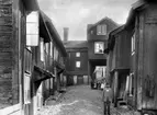 Tvåvånings bostadshus i trä.
Gårdsinteriör. Äldre man och 8 barn.
Reproduktion efter en bild från 1905.