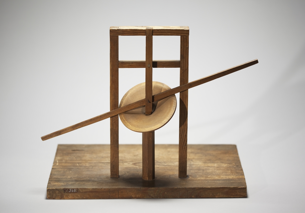 Modell ur Polhems mekaniska alfabet. Text på föremålet: "LXIV". Vid rotation av den lodräta axeln erhålles med hjälp av den vinkelställda skivan en vickande rörelse hos armen.