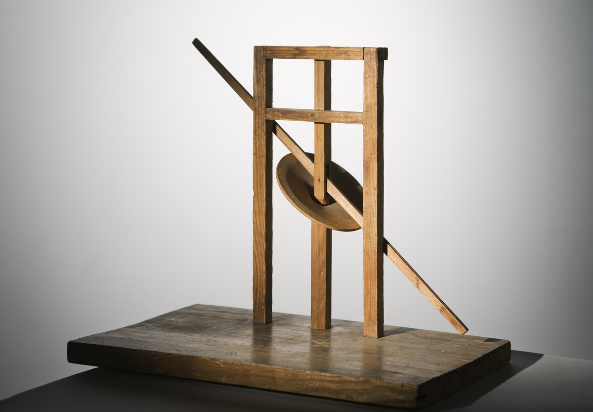 Modell ur Polhems mekaniska alfabet. Text på föremålet: "LXIV". Vid rotation av den lodräta axeln erhålles med hjälp av den vinkelställda skivan en vickande rörelse hos armen.