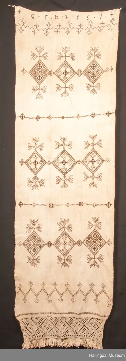 Tilnærmet rektangulært stk. hvit linlerret, håndvevet. Jarekanter i langsidene, faldet for hånd i ei kortside. Det vevde stykket ca. 130 cm langt og fortsetter i en knipling i firflettingsteknikk av renningstrådene. På linet er brodert mønster m/ 2-tråds, håndspunnet, mørk brunt ullgarn. Geometriske mønster i holbeinsting og korssting, nær falden er brodert 3 bokstaver (siste er D, to første er usikre) og årstallet 1757 i korssting og kjedessting.