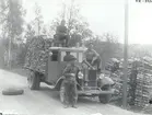 Vedtransport i trakten av Solstadström.  Stående på marken Gottfrid Andersson, på taket Erik Svensson, och på motorhuven Hjalmar Karlsson. De båda sistnämda var chaufförer. Reservhjulet är tillfälligt placerat på vägen, ringa trafik. och förvarades under körning alltid uppe på lasten.