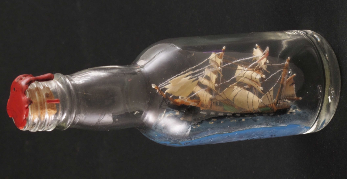 Miniatyr flaskeskute: Bark  Glass, klart. Kork, rød lakk. Innstøpt i bunnen:  MG (Moss Glassverk)  Sort skrog med hvitmalte "kanonporter". Blå sjø.  Utført av giver, hans nummer 84.