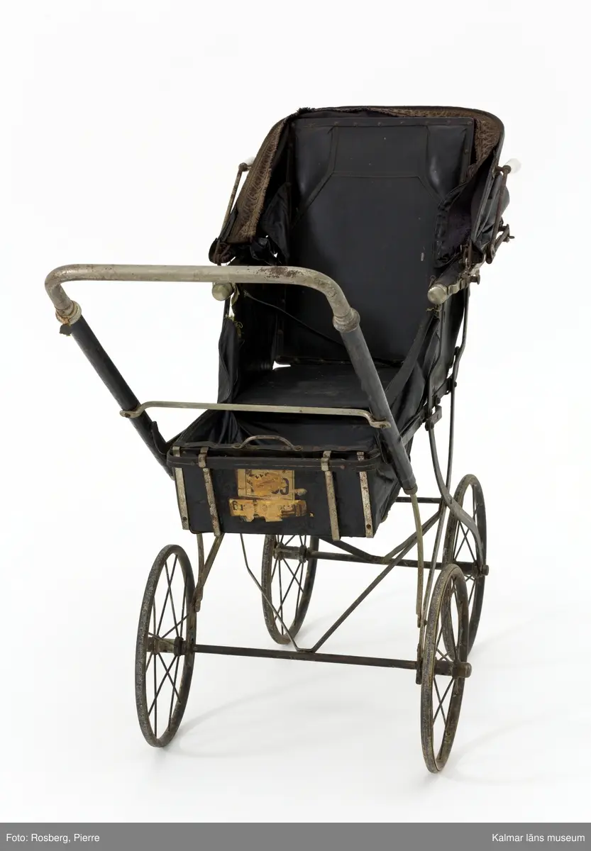 KLM 28494:121. Barnvagn, sittvagn. Stomme av metall, övervägande svartmålad med vita och gula dekorlinjer. Gummiklädda hjul. Sits av svart konstläder, delvis stoppad, med stomme av svartmålat trä. Konstlädret är fastnitat i stommen med nitar formade som små blommor. Sufflett av svart konstläder med handtag av vitt porslin. Sufflettens främre och bakre kant är försedd med vävt tygband av ylle i ursprungligen blå färg, numera blekt till en brun nyans. Baktill på vagnen en metallskylt: PHÖNIX. Framtill diverse fraktgodslappar, bl.a.: Charlottenberg.