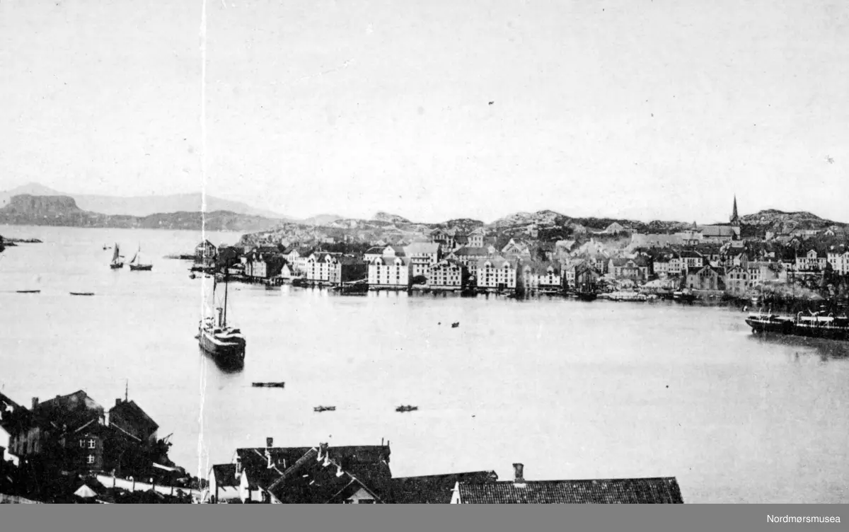 Havnebassenget sett fra Bjørnehaugen på Nordlandet rundt 1895. Her ser vi Kirkelandet med litt av Vågen og Johnsenhuken helt til høyre på andre siden av havnebasenget. Vi ser også Kirkelandet kirke som ruver på Lyhshaugen. Ellers så ser vi noe som kan likne på en molo fra Innlandet som stikker ut i bildet helt fra venstre, men denne er ikke med på kart 1905/1914. Mens helt i bakgrunnen ser vi Averøy kommune (1554) med Bremsneshatten. Ellers så ser vi to dampskip ute i havnebasenget. Fotograf er muligens Ranheimsæter, Ole Olsen. Kristiansunds historie nevner at det helt fra havnefogd Schjetnans dager, 1890-årene, hadde det vært et ønske om en molo i Sørsundet for å forbedre forholdene. Behovet for dette ble virkelig satt i fokus da saltskipet ";Norden"; fikk føle hvor vanskelig forholdene kunne være da det grunnstøtte i Lervika vinteren 1903. På foto KMb-2008-026.0045 kan en se noe som ligner på en fylling, og som sikkert er den samme som en kan se på KMb-1941-000.0200, men vet ikke noe om det er denne fyllingen som er forløperen til Sørsundmoloen. Den ble vel bygget på 1930-tallet. Dersom det finnes stoff fra Havnekommisjonens arkiv kan det antakelig være noe å finne der. Fra Nordmøre Museums fotosamlinger.
