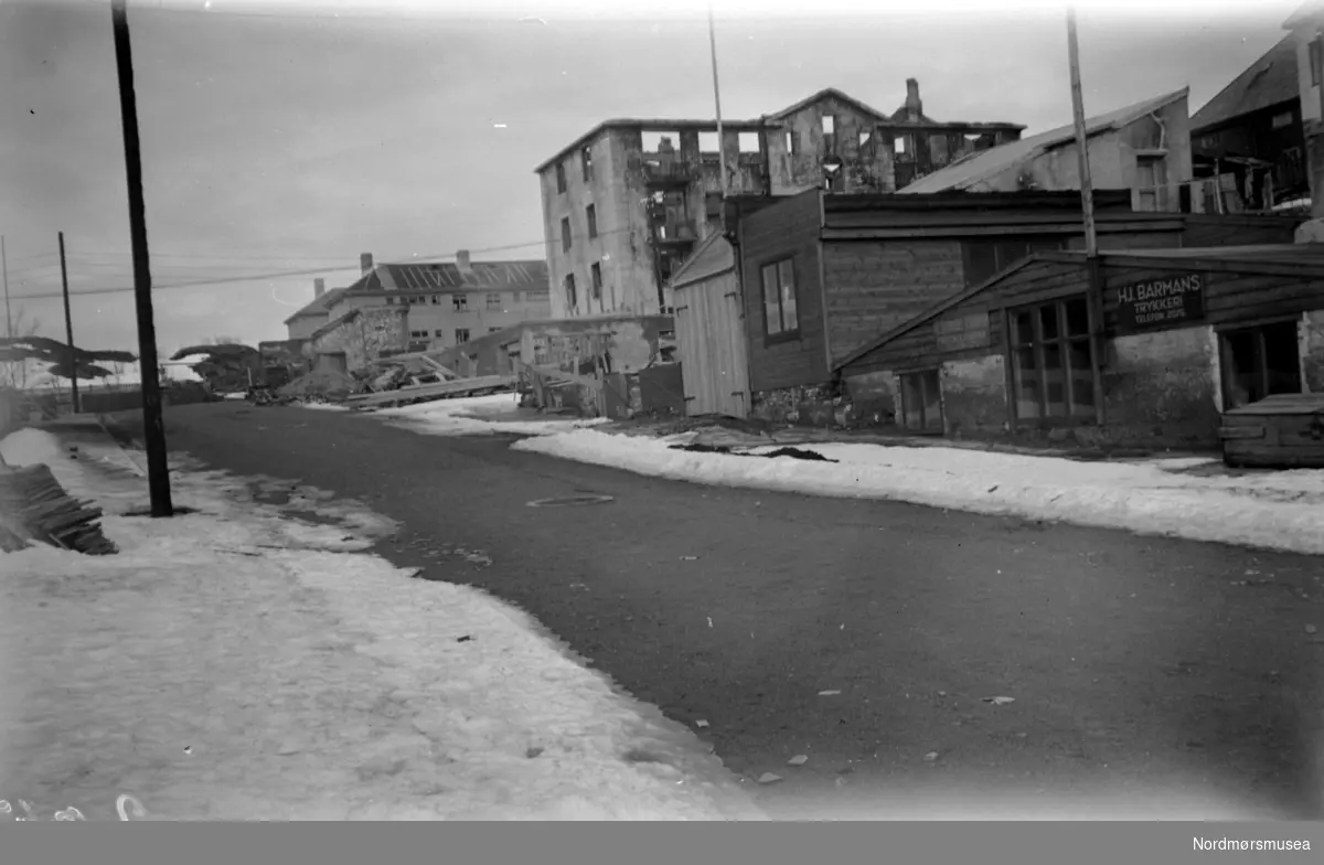 Gata til høyre for Festiviteten (Hans Neraas gate) i April 1941. Til høyre ser vi bokhandler Barmans trykkeri og bokbinderi (i brakker). Bak: Ruinene av ";Funksjonærboligen";. Lenger bak ser vi kommunale bolighus/leiegåder i Tollåsenga. Bildet er tatt i April 1941. Nordmøre Museum
