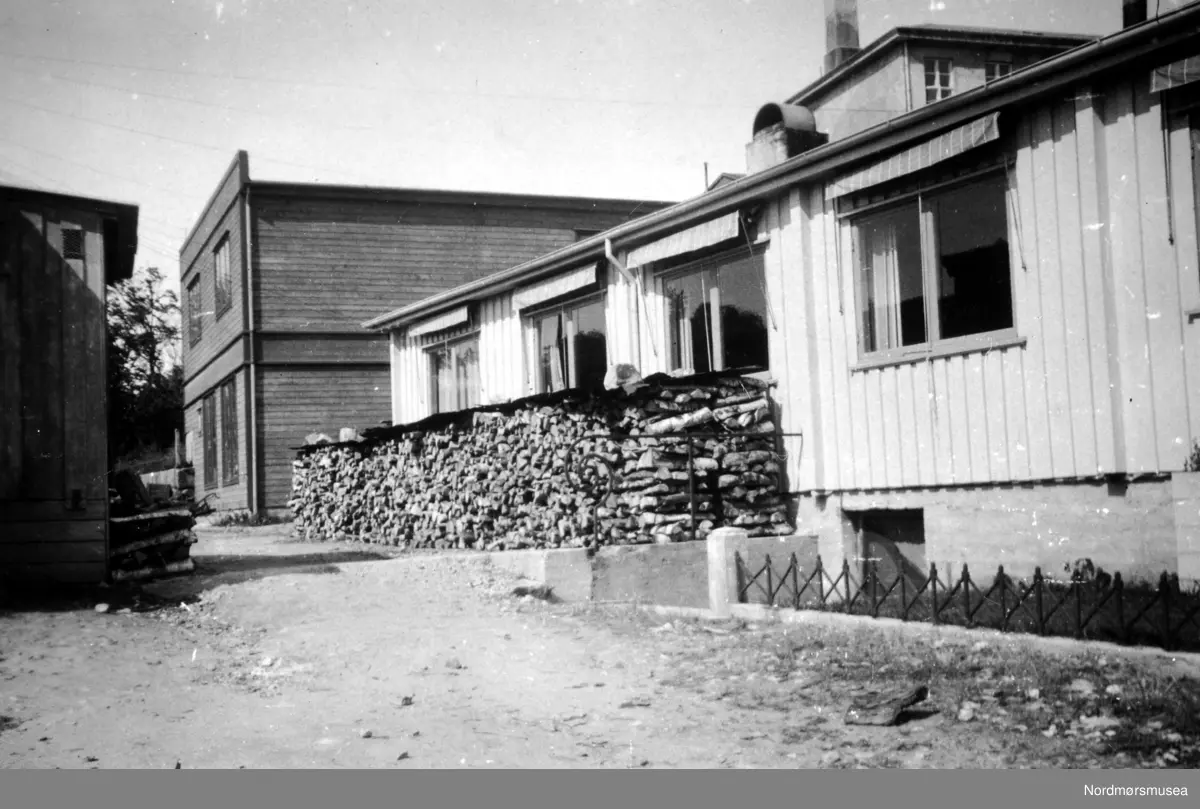 Utenfor Kristiansund og Nordmøres Forretningbanks brakke, juni 1943. Forsyning av vinterved stablet oppetter veggen. Brakka ligger her på Enggatens skole forhenværende tomt. I bakgrunnen til høyre ser vi taket til Kristiansund Offentlige Høgre Allmenskole (KOHAS), senere Langveien Ungdomsskole. Bildet er datert juni 1943. Nordmøre Museum.