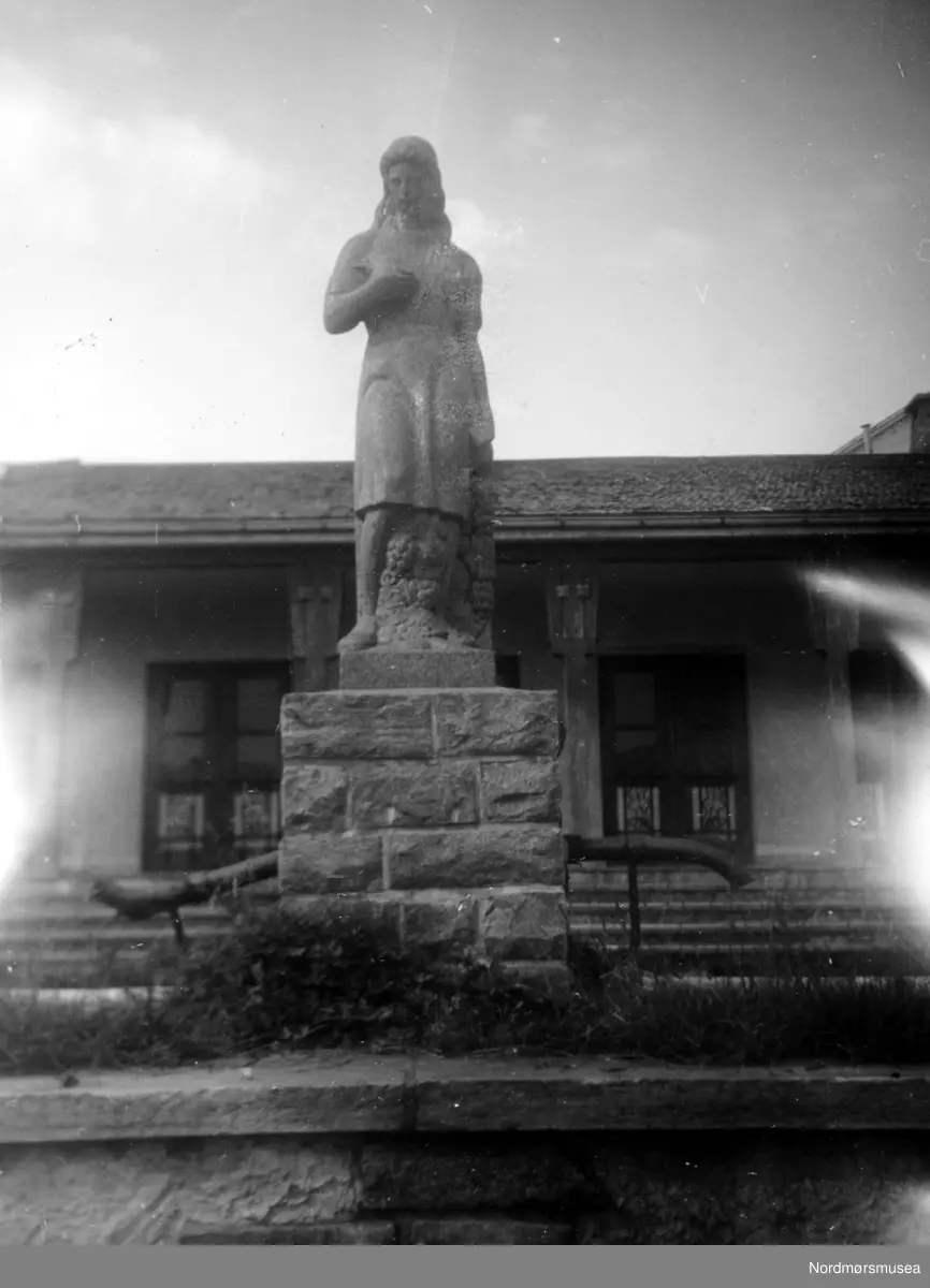 På bildet ser vi den nordre fløy av Soldatheimen med statuen i forgrunnen, i Lars Guttorms gate - nær Allanengen Skole. Statuen er utført av en tysk soldat ved navn Rainer, og forestiller en ung kvinne. Rainer, som var steinhugger og treskjærer i det sivile, laget statuen i en brakke i Vågen. Et vakkert kunstverk. Statuen er nå å finne i en privat hage i Kristiansund. Etter 1945 ble bygningen solgt til Kristiansund Arbeiderparti, som til per dags dato (august 1955) har brukt lokalet som samlingssted, møtelokale, samt til utleie for byens forskjellige foreninger. Det har også blitt brukt til offentlig valglokale i den store salen. I disse dager (per august 1955) holder de på å rive Soldatheimen, for å gi plass til byggetomter.
Kilde (blant andre): Tidens Krav, Fredag 14. september 2007, side 20
Bildet er fra Nordmøre Museums fotosamlinger.